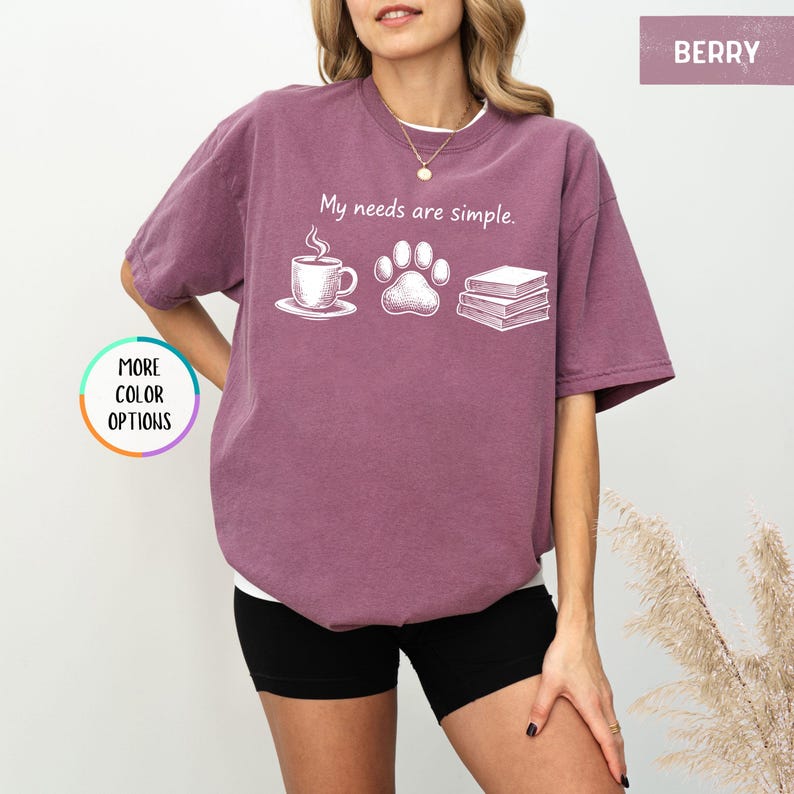 May include: Berry-colored t-shirt with the phrase "My needs are simple." The graphic includes a coffee cup, a paw print, and a stack of books. The shirt is a relaxed fit.