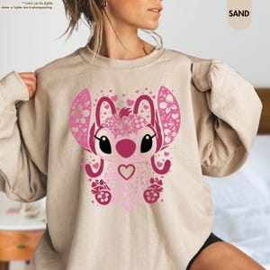 May include: Sand-colored sweatshirt featuring a pink cartoon character design. The character has large ears, a heart-shaped nose, and a heart on its chest. The design incorporates floral and heart patterns.