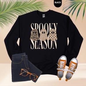 May include: A black long-sleeve sweatshirt featuring the text SPOOKY SEASON in a cream font. Three ghost illustrations are centered between the words, each with a different pattern: camouflage, leopard print, and a brown and cream checkerboard.