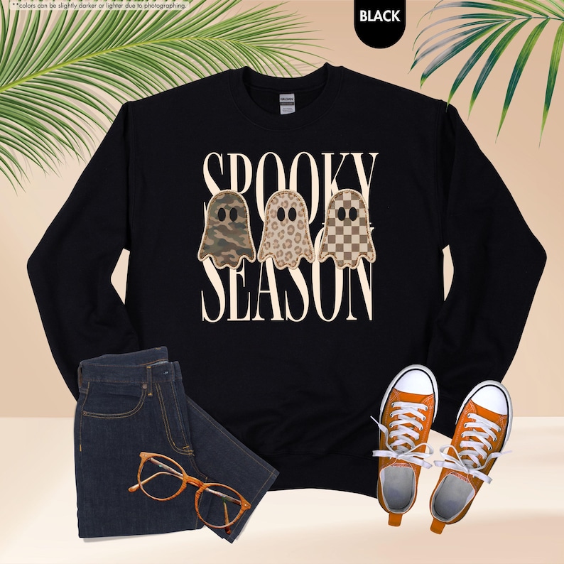 May include: A black long-sleeve sweatshirt featuring the text SPOOKY SEASON in a cream font. Three ghost illustrations are centered between the words, each with a different pattern: camouflage, leopard print, and a brown and cream checkerboard.
