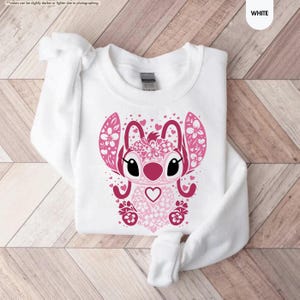 May include: White sweatshirt featuring a pink graphic of a cartoon character with large ears, floral accents, and heart details. The design is centered on the front of the long-sleeved top. The word "WHITE" is in the upper right corner.
