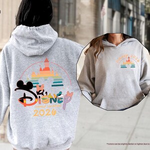 May include: A light gray hoodie with a front pocket and a hood. The back features a colorful Disney graphic with a castle, Mickey Mouse ears, and the text "Disney Summer Vacation 2026". The front has a smaller matching graphic.