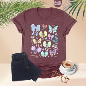 May include: Maroon heather t-shirt with a graphic of colorful bows and princess names. The image includes dark wash jeans, a coffee cup with a heart design, and spectacles.