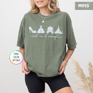 May include: Moss green t-shirt featuring a whimsical illustration of a glass slipper, ball gown, carriage, and castle. The words "meet me at midnight" are written in a script font below the illustrations. The shirt has short sleeves and a relaxed fit.