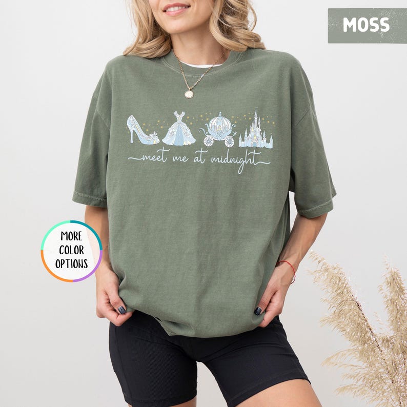May include: Moss green t-shirt featuring a whimsical illustration of a glass slipper, ball gown, carriage, and castle. The words "meet me at midnight" are written in a script font below the illustrations. The shirt has short sleeves and a relaxed fit.