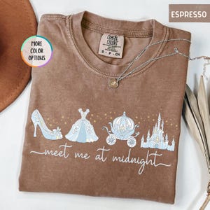 May include: Espresso-colored t-shirt with a Cinderella-themed graphic. The design includes a high heel, ball gown, carriage, and castle, with the words "meet me at midnight." A silver necklace is draped across the shirt.