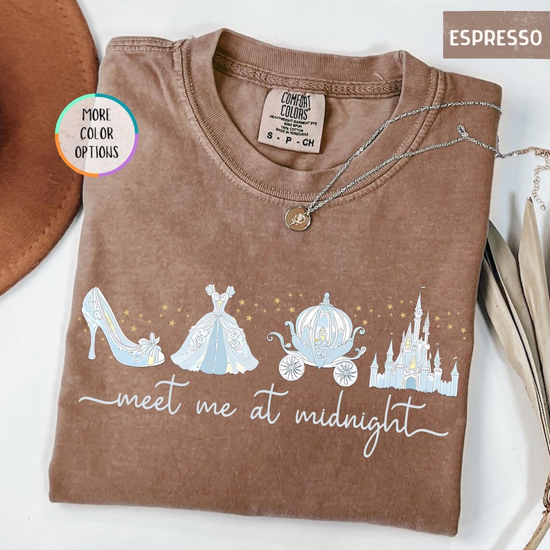 May include: Espresso-colored t-shirt with a Cinderella-themed graphic. The design includes a high heel, ball gown, carriage, and castle, with the words "meet me at midnight." A silver necklace is draped across the shirt.