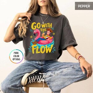 Puede incluir: Una camiseta de manga corta color gris carb&oacute;n con un gr&aacute;fico de un vaquero sentado en un flotador de flamenco rosa sobre agua azul. El texto "GO WITH THE FLOW" est&aacute; impreso en amarillo encima y debajo del gr&aacute;fico. El color es "PEPPER".