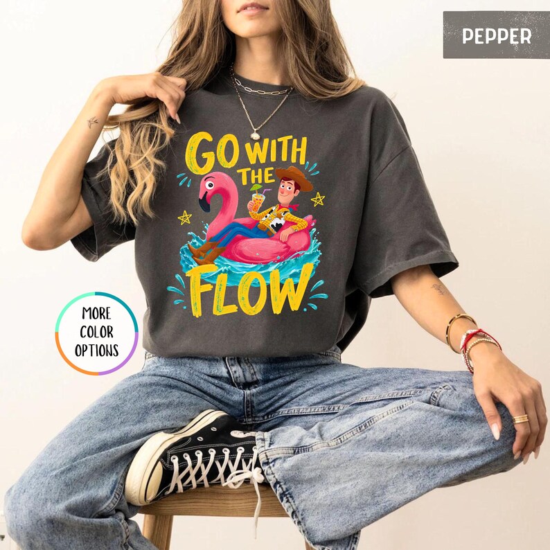 Puede incluir: Una camiseta de manga corta color gris carb&oacute;n con un gr&aacute;fico de un vaquero sentado en un flotador de flamenco rosa sobre agua azul. El texto "GO WITH THE FLOW" est&aacute; impreso en amarillo encima y debajo del gr&aacute;fico. El color es "PEPPER".