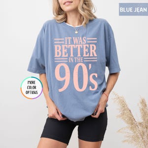 May include: A blue jean colored short-sleeve t-shirt featuring the light pink text IT WAS BETTER IN THE 90's printed on the front. The shirt has a crew neckline and a relaxed fit. A circular graphic on the left side displays the text MORE COLOR OPTIONS.