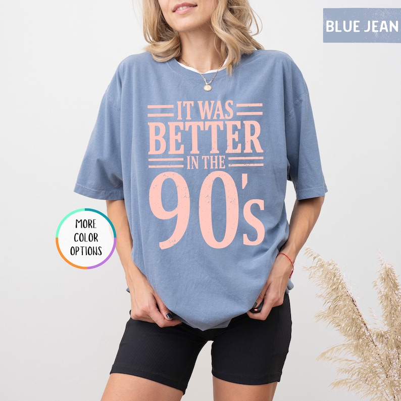 May include: A blue jean colored short-sleeve t-shirt featuring the light pink text IT WAS BETTER IN THE 90's printed on the front. The shirt has a crew neckline and a relaxed fit. A circular graphic on the left side displays the text MORE COLOR OPTIONS.