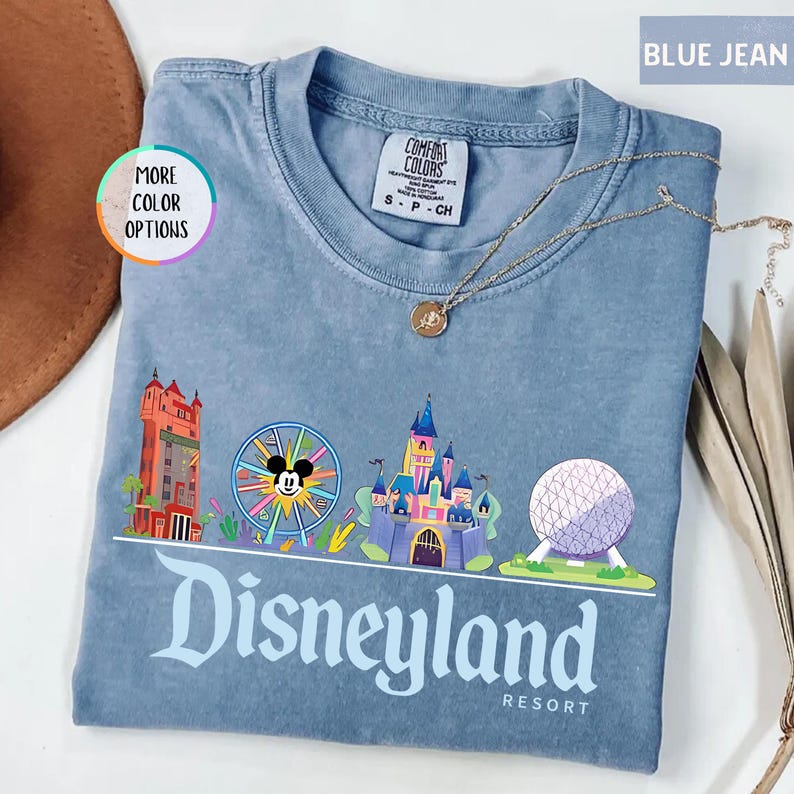 Op de afbeelding: Blauw spijkerbroekkleurig T-shirt met een Disneyland Resort-afbeelding. De afbeelding bevat een reuzenrad met Mickey Mouse, een kasteel en de tekst "Disneyland Resort". Een gouden ketting ligt over het shirt.