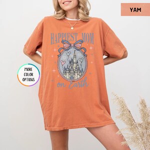 May include: Orange t-shirt with the text "Happiest Mom on Earth" in light blue. The design includes a castle illustration within a decorative oval frame, with a pink and blue bow at the top. The shirt has short sleeves.