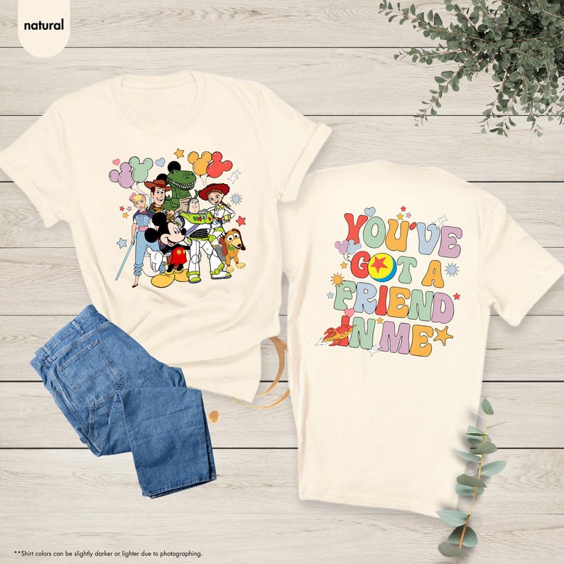 May include: Cream-colored t-shirt featuring a colorful graphic of Disney characters, including Mickey Mouse, Woody, and Buzz Lightyear. The back of the shirt displays the text "YOU'VE GOT A FRIEND IN ME" in a retro font. Paired with blue denim jeans.