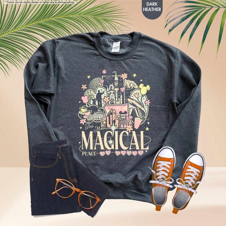 May include: Dark heather gray sweatshirt with a colorful graphic that reads "The Most Magical Place." The design includes a castle, Mickey Mouse ears, and other theme park elements. The image also shows dark wash jeans, orange sneakers, and eyeglasses.