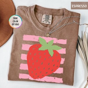May include: An espresso-colored short-sleeve t-shirt featuring a large red strawberry graphic with a green stem, layered over horizontal light pink stripes. The shirt has a crew neckline and a Comfort Colors brand label inside the collar.