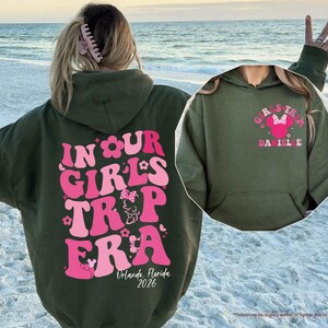 Peut inclure: Un sweat &agrave; capuche vert olive avec un motif rose et blanc au dos indiquant "IN OUR GIRLS TRIP ERA" avec "Orlando, Florida 2026" en dessous. Un motif plus petit sur le devant indique "GIRLS TRIP" avec un design Minnie Mouse.