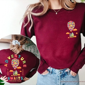 Puede incluir: Sudadera granate con un gr&aacute;fico de globo aerost&aacute;tico y casa coloridos. La parte posterior de la sudadera presenta el texto "ADVENTURE AWAITS PARADISE FALLS" en un arco iris sobre el mismo gr&aacute;fico. La sudadera se usa con jeans azules.
