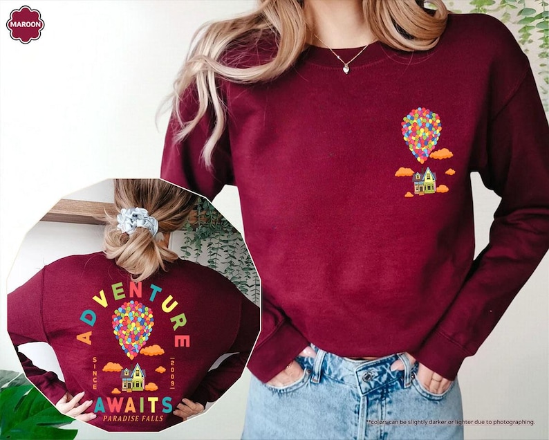 Puede incluir: Sudadera granate con un gr&aacute;fico de globo aerost&aacute;tico y casa coloridos. La parte posterior de la sudadera presenta el texto "ADVENTURE AWAITS PARADISE FALLS" en un arco iris sobre el mismo gr&aacute;fico. La sudadera se usa con jeans azules.