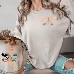 May include: Sand-colored sweatshirt with a Disney 2020 graphic. The design features a castle, rainbow, and text. The sweatshirt is a neutral color and has a crew neck.