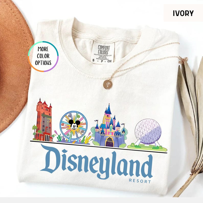 Op de afbeelding: Ivoor kleurig T-shirt met een Disneyland Resort afbeelding. Het ontwerp bevat iconische park bezienswaardigheden zoals een kasteel, een Mickey Mouse reuzenrad en een futuristische bol. Het woord "Disneyland" is in blauw geschreven.
