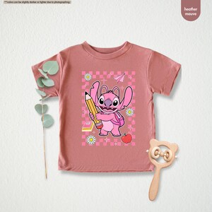 May include: Heather mauve t-shirt with a pink cartoon character holding a pencil, backpack, apple, and daisies. The design is set against a pink and white checkered background. The shirt is a short-sleeved crew neck.
