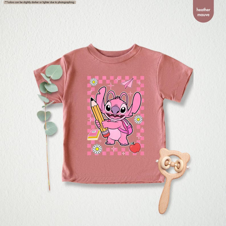May include: Heather mauve t-shirt with a pink cartoon character holding a pencil, backpack, apple, and daisies. The design is set against a pink and white checkered background. The shirt is a short-sleeved crew neck.