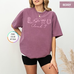 May include: Berry-colored t-shirt with a white undershirt. The shirt features a light pink graphic of a cowboy boot, hat, bow, horseshoe, and the words "Cloud 9". The shirt is a relaxed fit.