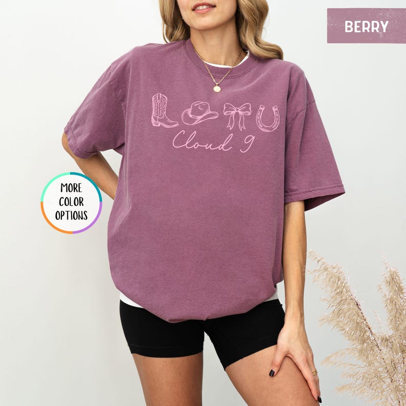 May include: Berry-colored t-shirt with a white undershirt. The shirt features a light pink graphic of a cowboy boot, hat, bow, horseshoe, and the words "Cloud 9". The shirt is a relaxed fit.