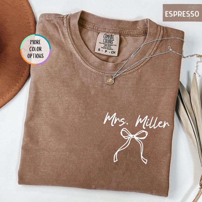 Puede incluir: Camiseta Comfort Colors color espresso con el nombre "Mrs. Miller" y un lazo blanco. Un collar plateado con un colgante est&aacute; drapeado sobre la camiseta. La camiseta tiene cuello redondo y mangas cortas. Las palabras "More Color Options" est&aacute;n en un c&iacute;rculo.