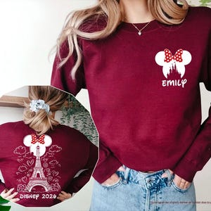 May include: A maroon long-sleeve sweatshirt featuring a white Minnie Mouse silhouette with a red polka dot bow and the name EMILY on the front. The back displays a white graphic of the Eiffel Tower, clouds, hearts, and DISNEY 2026 text.