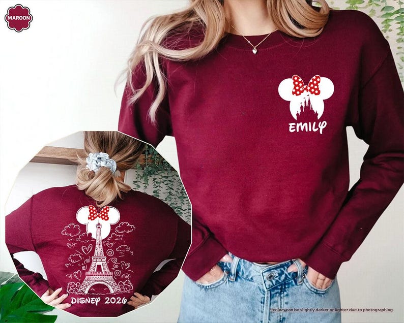 May include: A maroon long-sleeve sweatshirt featuring a white Minnie Mouse silhouette with a red polka dot bow and the name EMILY on the front. The back displays a white graphic of the Eiffel Tower, clouds, hearts, and DISNEY 2026 text.