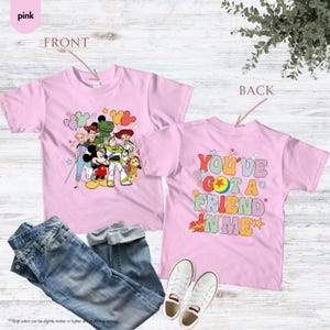 May include: Pink t-shirts with a graphic of Disney characters on the front and the text "YOU'VE GOT A FRIEND IN ME" on the back. The shirts are displayed with blue jeans and white sneakers.