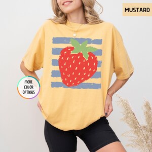 May include: A mustard yellow short-sleeve t-shirt featuring a graphic of a red strawberry with green leaves centered over horizontal blue stripes. The text MUSTARD appears in the top right corner, and a circular badge reads MORE COLOR OPTIONS.
