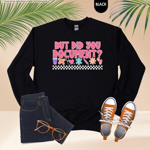 Puede incluir: Sudadera negra con el texto "BUT DID YOU DOCUMENT?" en rosa y blanco, junto con gr&aacute;ficos m&eacute;dicos. La imagen tambi&eacute;n muestra unos vaqueros azules, gafas naranjas y zapatillas deportivas naranjas.