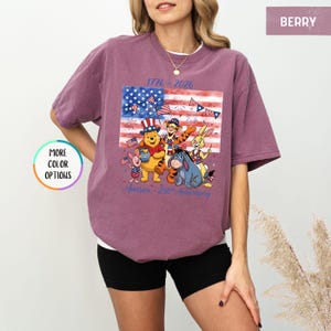 May include: Berry-colored t-shirt featuring a graphic of Winnie the Pooh and friends in front of an American flag. The shirt has the text "1776-2026" and "America - 250th Anniversary" in blue. The shirt is a short-sleeved, crew neck style.