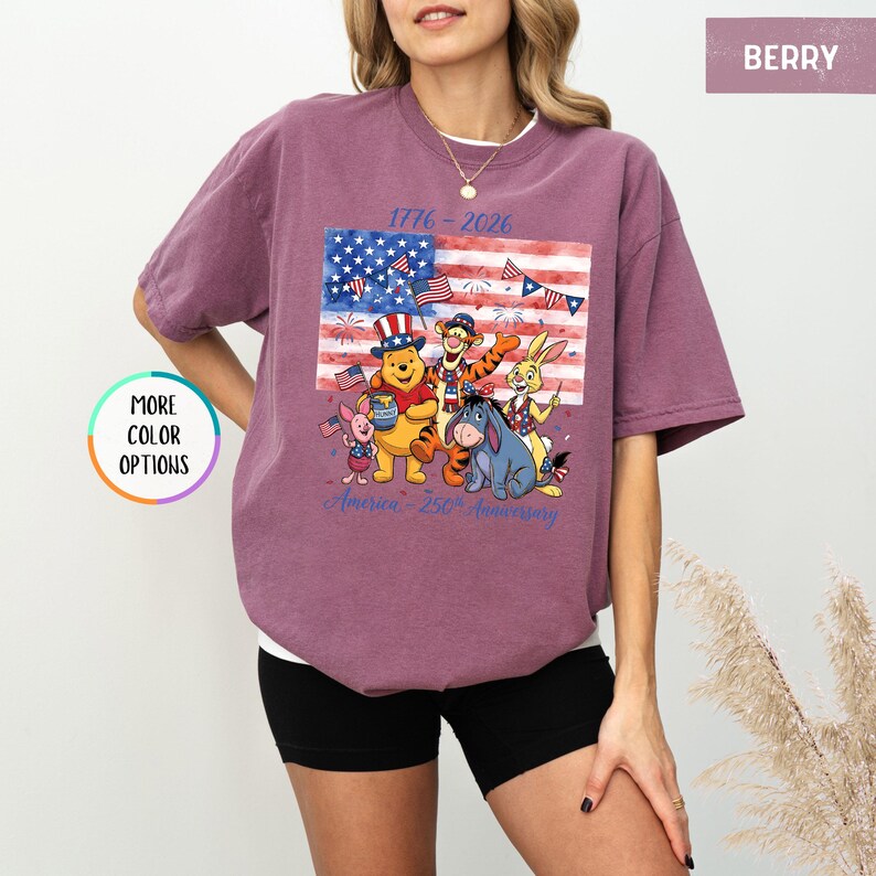 May include: Berry-colored t-shirt featuring a graphic of Winnie the Pooh and friends in front of an American flag. The shirt has the text "1776-2026" and "America - 250th Anniversary" in blue. The shirt is a short-sleeved, crew neck style.