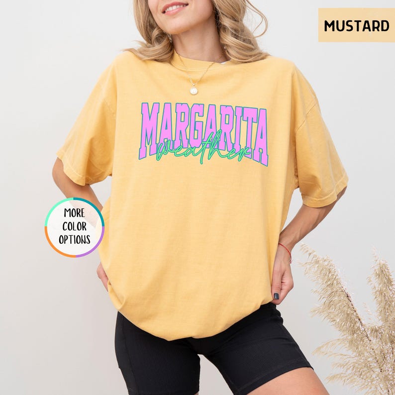 May include: Mustard-colored t-shirt with the text "MARGARITA weather" in a layered design of pink and green. The shirt is a relaxed fit and has short sleeves. The image also shows a "More Color Options" button.