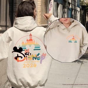 May include: Sand-colored hoodie with a Disney graphic on the back, featuring Mickey Mouse, Minnie Mouse, and a castle design. The text "Disney Family Vacation 2026" is also visible. A smaller graphic is on the front. The hoodie has a drawstring hood.