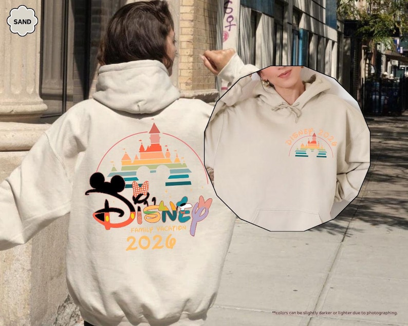 May include: Sand-colored hoodie with a Disney graphic on the back, featuring Mickey Mouse, Minnie Mouse, and a castle design. The text "Disney Family Vacation 2026" is also visible. A smaller graphic is on the front. The hoodie has a drawstring hood.