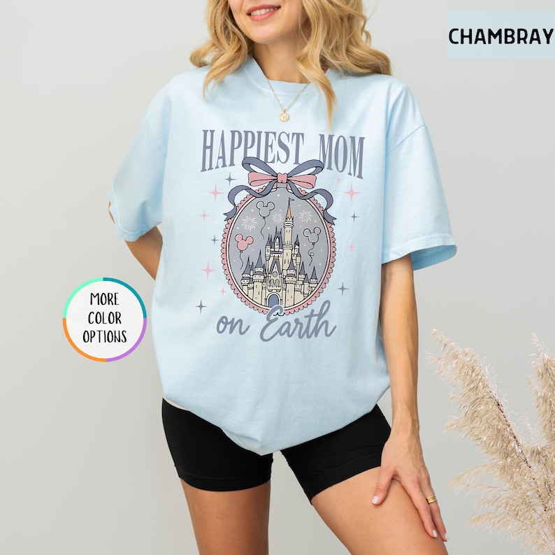 May include: Light blue t-shirt with the text "Happiest Mom on Earth" and a castle graphic. The graphic includes a pink bow, Mickey Mouse balloons, and a castle design. The shirt has short sleeves and a crew neck.