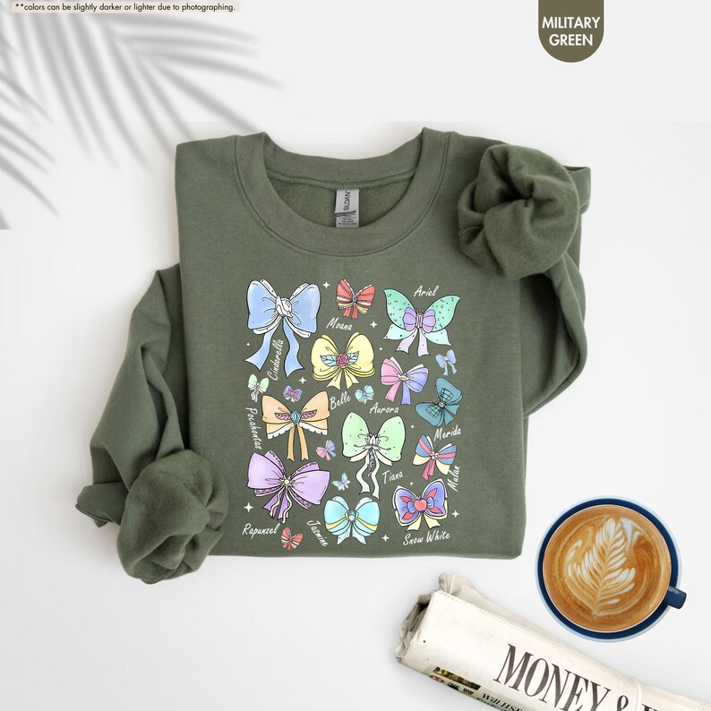 May include: Military green sweatshirt with a colourful bow design featuring princess names such as Ariel, Belle, and Cinderella. The design includes butterflies and is printed on a crew neck sweatshirt. A cup of coffee and a newspaper are in the image.