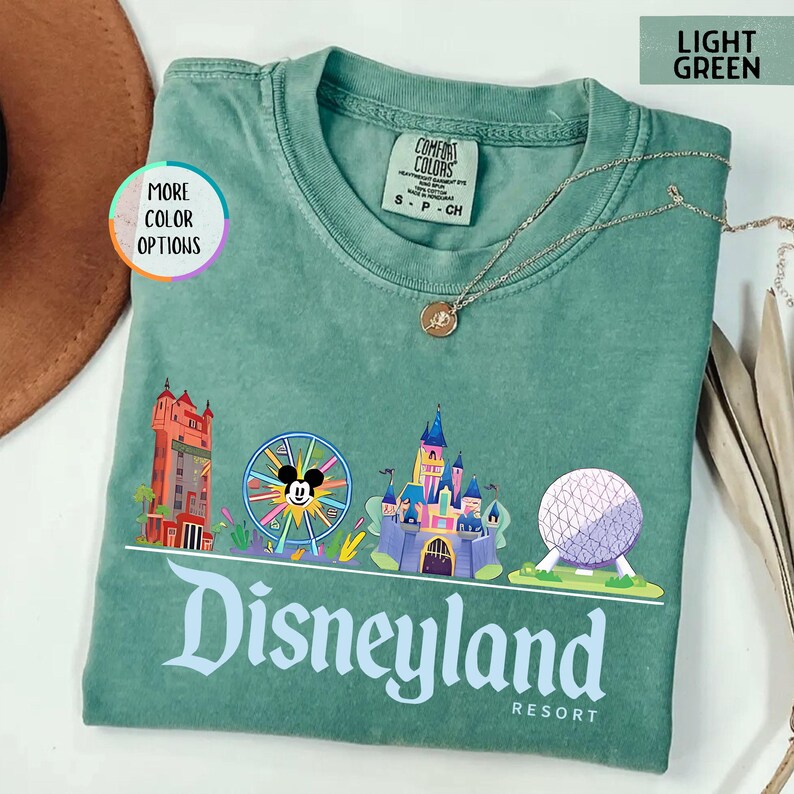 Op de afbeelding: Lichtgroen T-shirt met een Disneyland Resort-afbeelding. Het ontwerp bevat illustraties van iconische parklandmarks, waaronder een kasteel, een reuzenrad met Mickey Mouse en Spaceship Earth. De tekst "Disneyland Resort" staat onderaan afgedrukt.