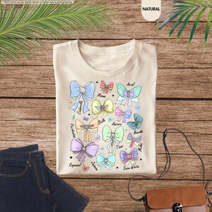 May include: A cream-coloured t-shirt with a colourful bow design featuring character names such as Cinderella, Ariel, and Snow White. The bows are in various colours, including blue, pink, and green. The shirt is on a wooden surface.