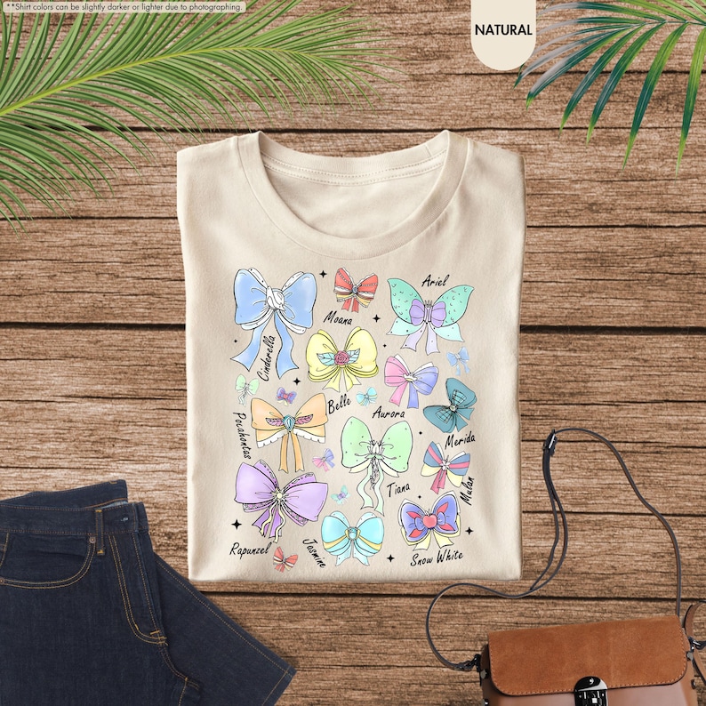 May include: A cream-coloured t-shirt with a colourful bow design featuring character names such as Cinderella, Ariel, and Snow White. The bows are in various colours, including blue, pink, and green. The shirt is on a wooden surface.