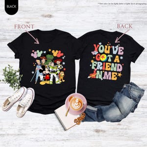 May include: Black t-shirts with a colorful graphic of Disney characters on the front and the phrase "YOU'VE GOT A FRIEND IN ME" on the back. The shirts are displayed with jeans, shoes, and a coffee cup.