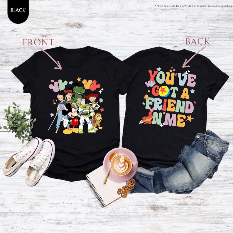 May include: Black t-shirts with a colorful graphic of Disney characters on the front and the phrase "YOU'VE GOT A FRIEND IN ME" on the back. The shirts are displayed with jeans, shoes, and a coffee cup.