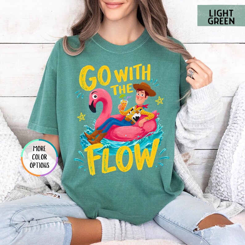 Puede incluir: Una camiseta verde claro con un gr&aacute;fico de un vaquero de dibujos animados sentado en un flotador de flamenco rosa en agua azul. El texto GO WITH THE FLOW aparece en letras amarillas. Incluye una insignia circular que dice MORE COLOR OPTIONS.