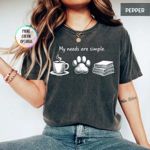 May include: Pepper-colored t-shirt with the phrase "My needs are simple." and illustrations of a coffee cup, paw print, and books. The shirt has a crew neck and short sleeves. The words "More Color Options" are in a circle on the left.