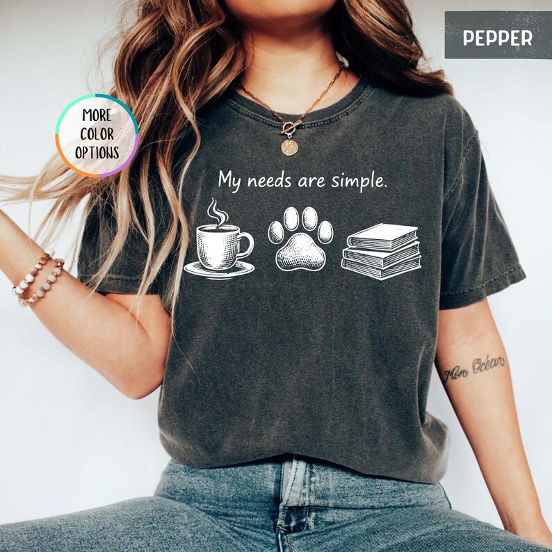 May include: Pepper-colored t-shirt with the phrase "My needs are simple." and illustrations of a coffee cup, paw print, and books. The shirt has a crew neck and short sleeves. The words "More Color Options" are in a circle on the left.